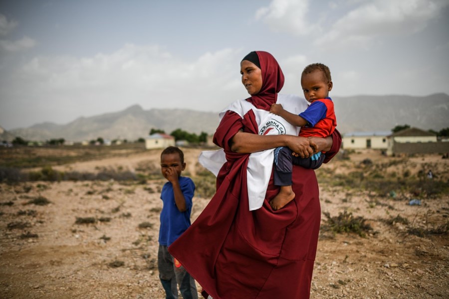 En kvinne med Røde Kors-vest står ute på en åpen slette med et lite barn i armene.