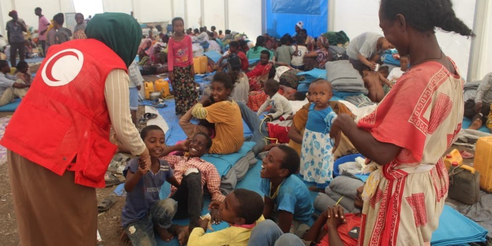Mange mennesker, spesielt små barn, samlet i et stort telt. En kvinne fra Sudan Røde Halvmåne snakker med noen små barn.