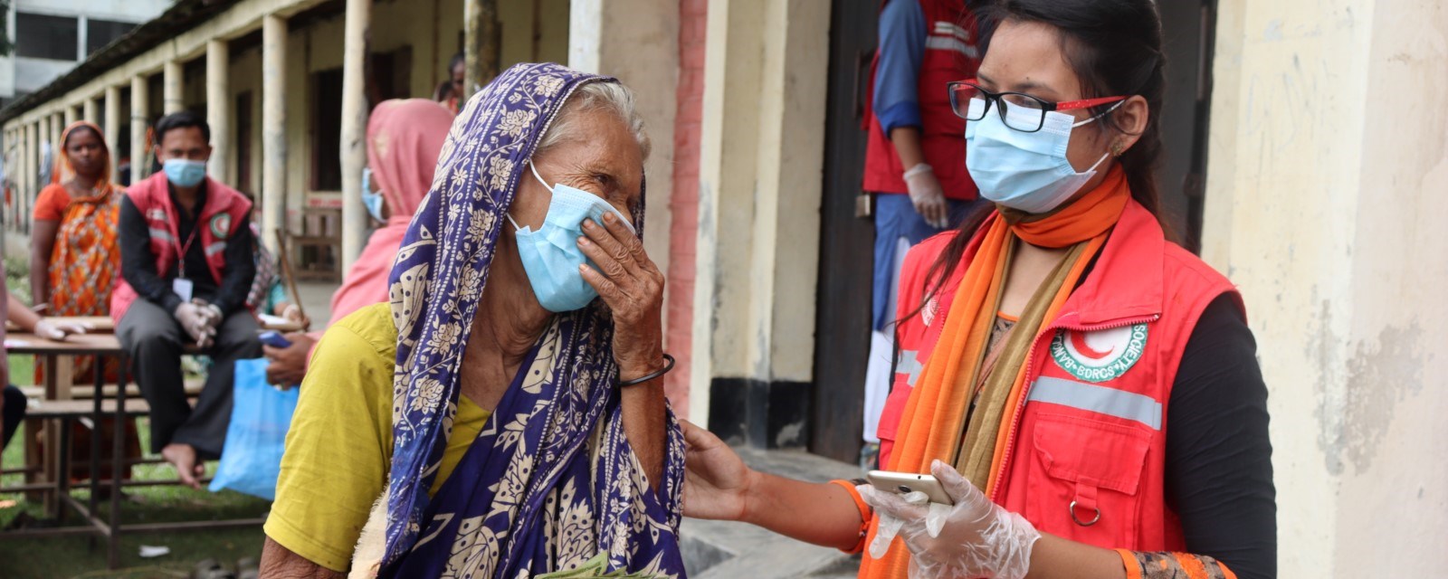 En godt voksen kvinne med munnbind ser på en kvinnelig frivillig fra Bangladesh Røde Halvmåne.