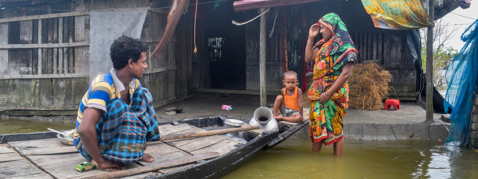 En mann og et lite barn sitter i en båt som ligger utenfor et hus. En kvinne står i vannet.