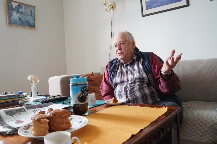 En eldre mann sitter i en sofa og snakker med kanelbolle og kaffe på bordet