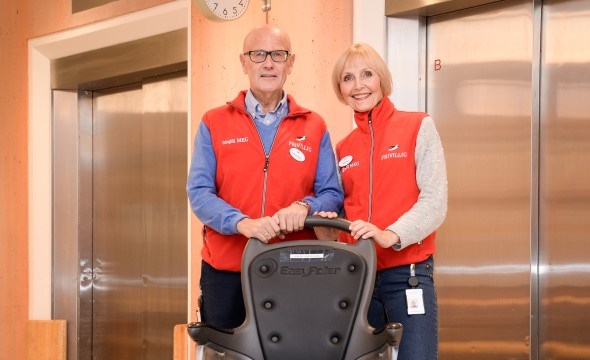 Bildet viser to frivillige i røde vester som triller en tom rullestol for å illustrere hvordan de kan hjelpe pasienter ved sykehuset.
