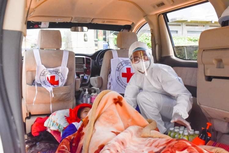 En kvinne i smittevarnsutstyr behandler en pasient i en Røde Kors-bil.
