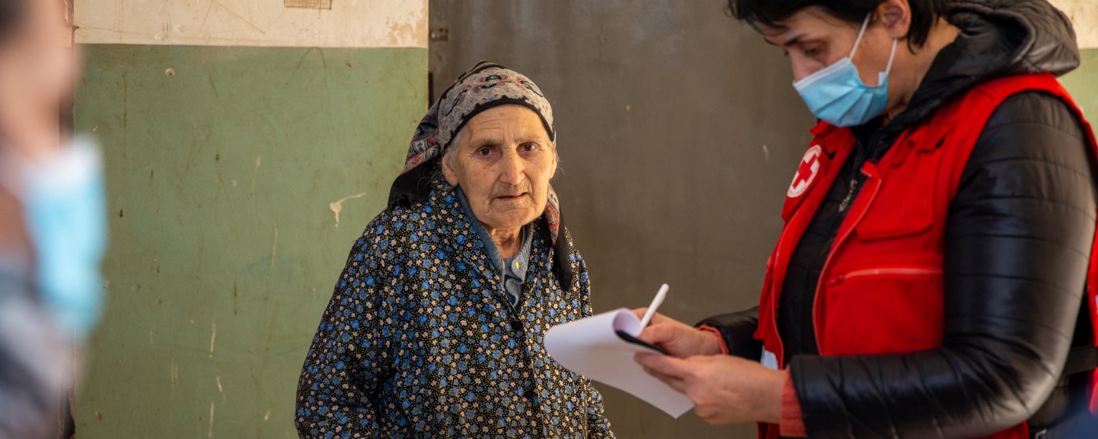 En eldre kvinne ser inn i kamera mens en frivillig fra Røde Kors står ved siden av og skriver på et papir.
