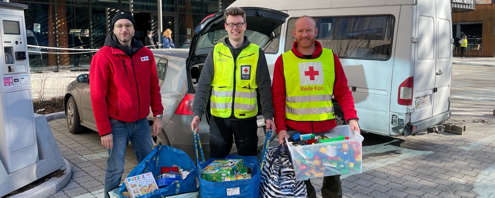 Tre menn fra Røde Kors står utenfor et bygning med store bæreposer med leker