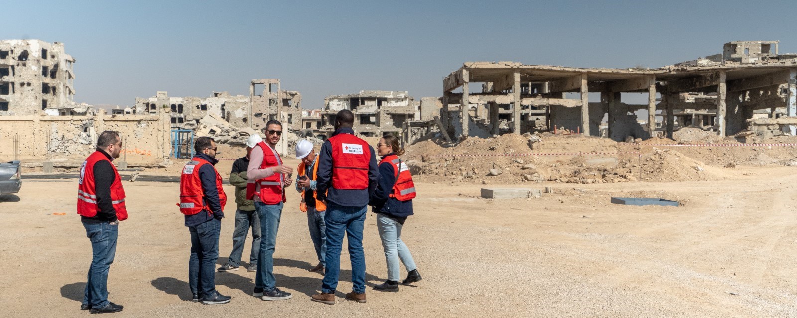 Åtte mennesker fra Røde Kors står foran flere utbombede bygninger 