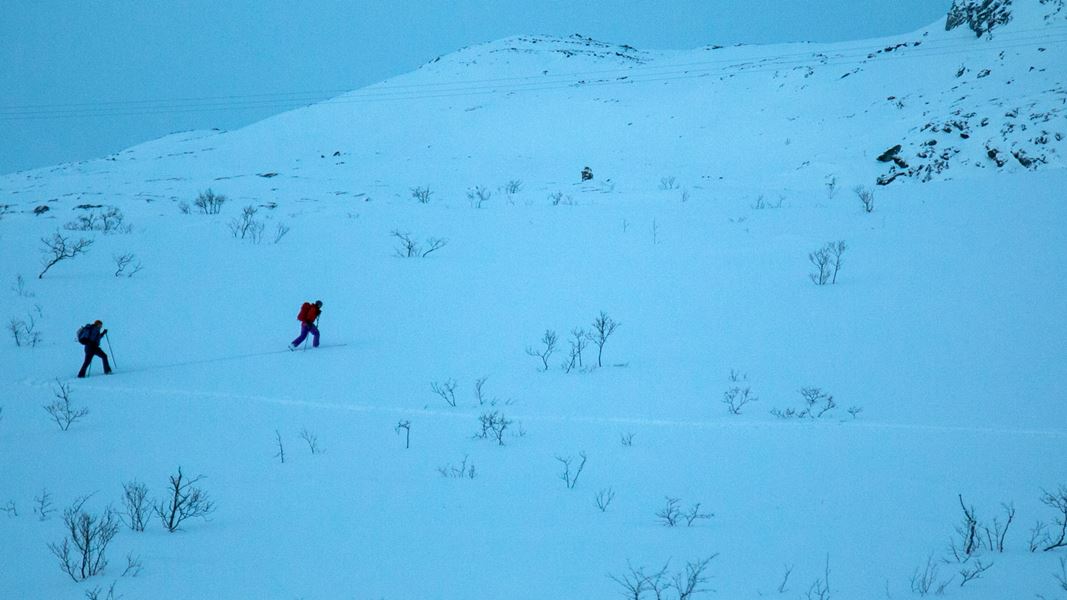 to menensker på vei opp på ski i slakk snødekket fjellside i blålysningen