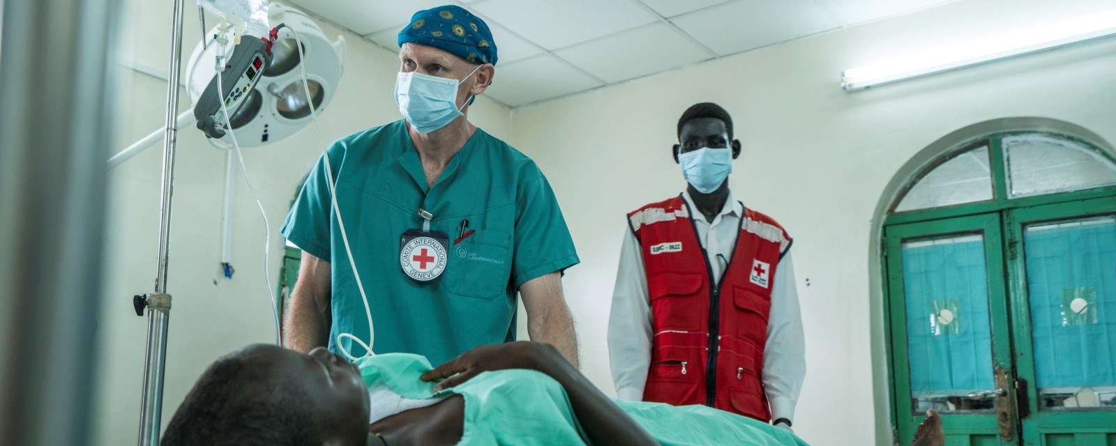 En norsk lege ikledd turkise operasjonsklær står sammen med en lokal sørsudansk frivillig i rød vest, over et operasjonsbord med en pasient liggende klar for behandling