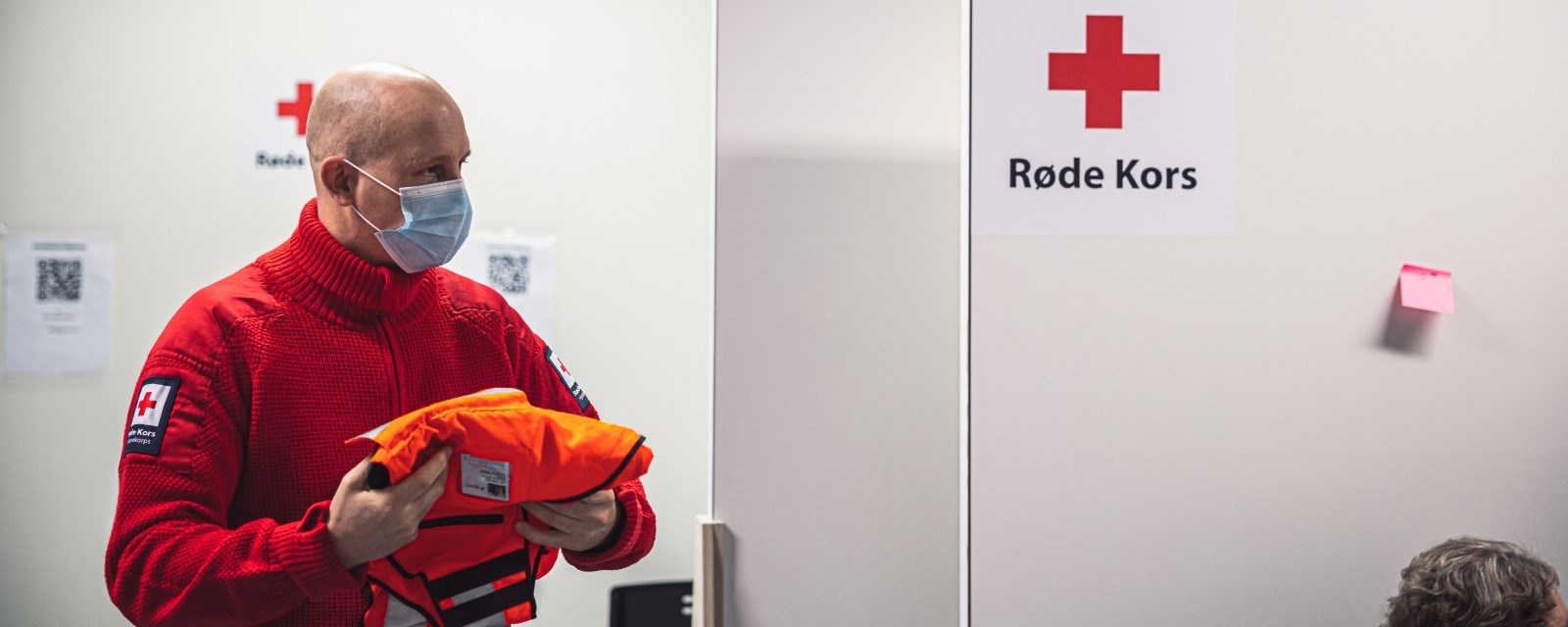 En mann med Røde Kors-henser og munnbind pakker sammen en oransje vest.