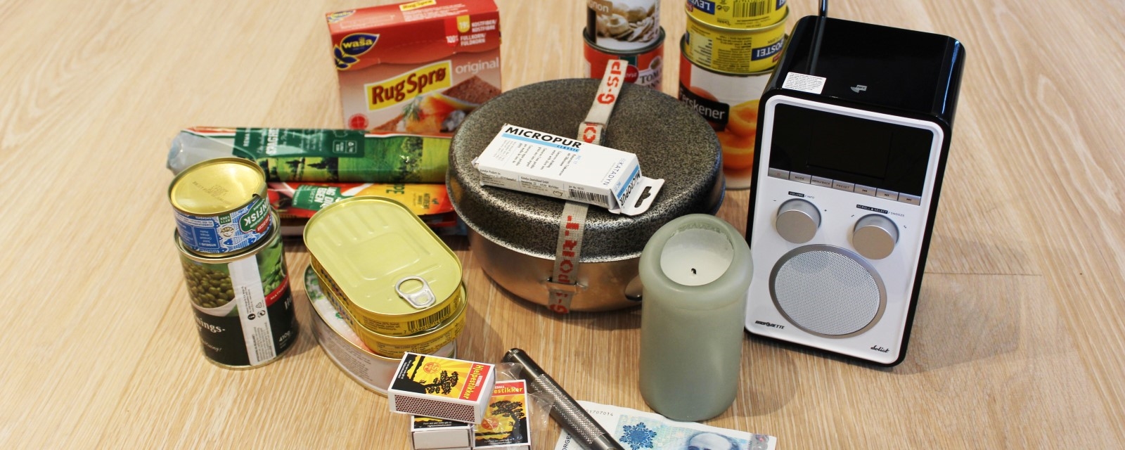 På bildet er diverse hermetikk stilt opp, et stormkjøkken, vannrensetabletter, fyrstikker, lommelykt, batterier, radio, stearinlys og litt kontanter