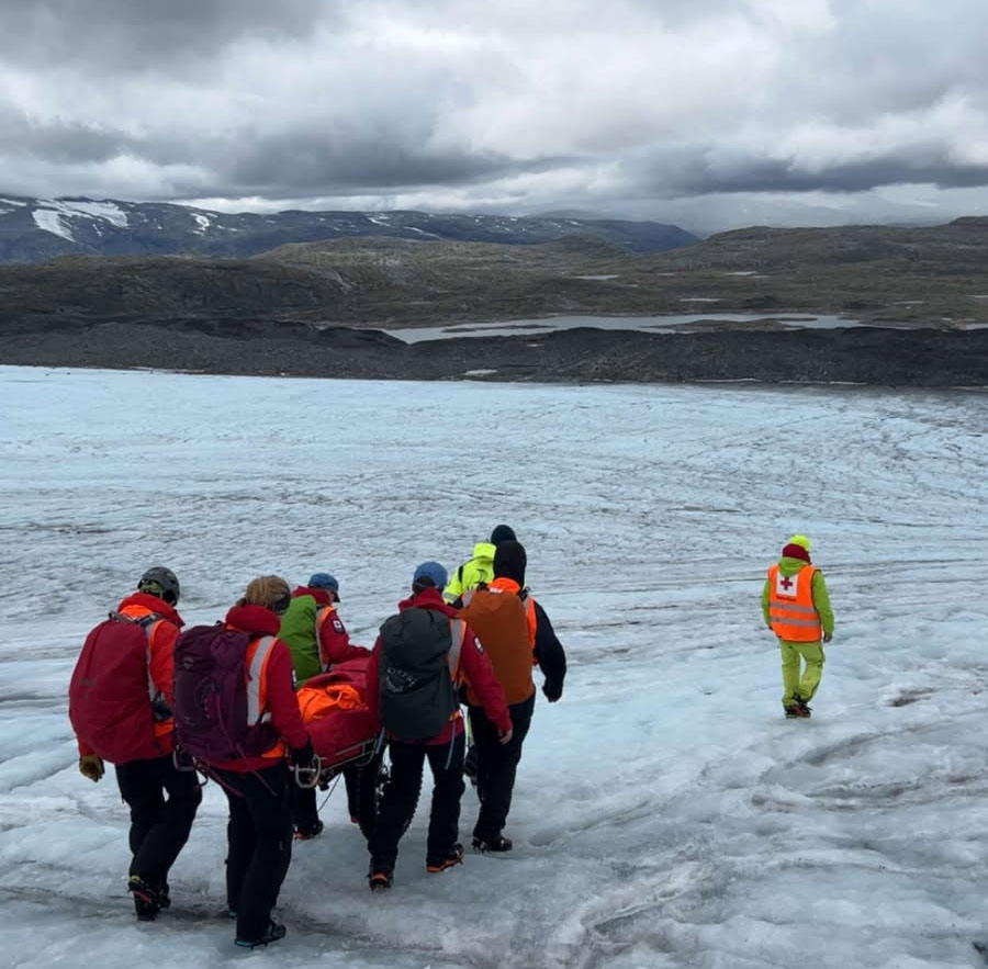 Flere frivillige fra hjelpekorpset bærer en skadet person i fjellet, på snøen.