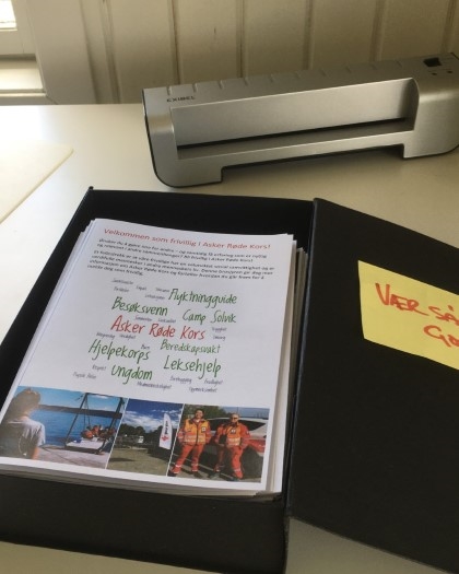 En boks full av rekrutteringsbrosjyrer for Asker Røde Kors som ligger på et bord.