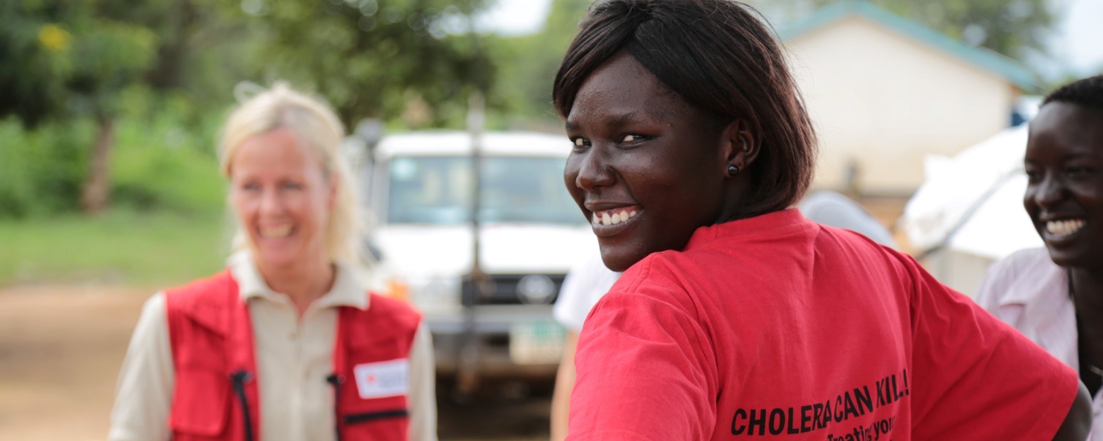 En sør-sudansk kvinner snur seg og smiler til kamera ute i felten. På T-skjorten står det "kolera kan ta liv"