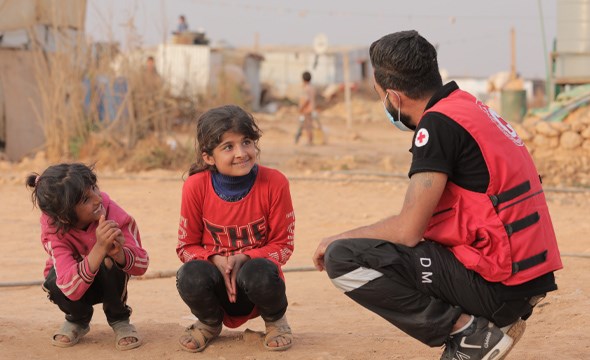 Frivillig på huk med to barn foran flyktningleir