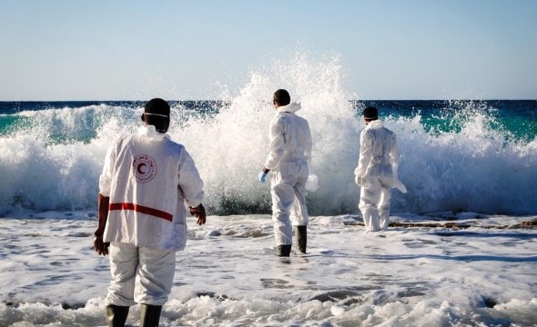 Tre personer i hvite arbeidsdresser og gummistøvler går ut mot havet