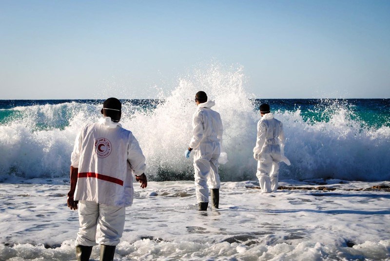 Tre personer i hvite arbeidsdresser og gummistøvler går ut mot havet