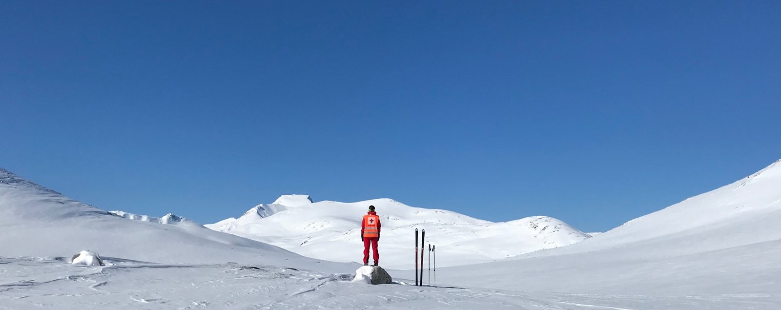 Bildet viser en person i Røde Kors-uniform som står ved siden av et par ski, omgitt av blå himmel og snødekte fjell.
