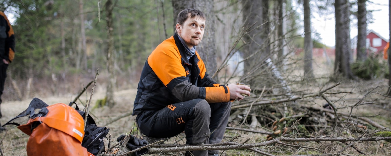 Terje sitter på en trestamme og spiser lunsj i fullt skogsarbeider-hyre. Ved siden av står termos, hjelm og motorsag