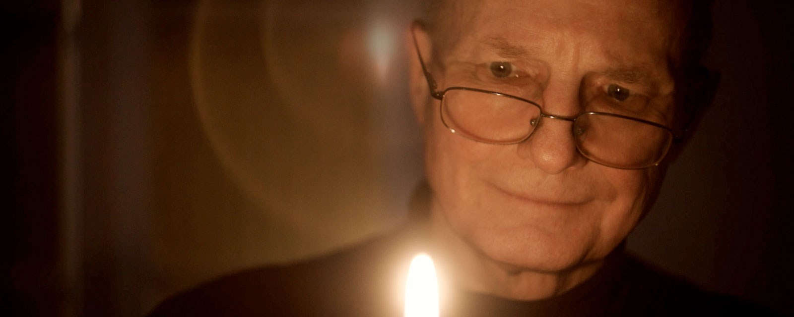 En eldre mann tenner et stearinlys i et ellers mørkt rom. I konturene i ansiktet aner vi et smil