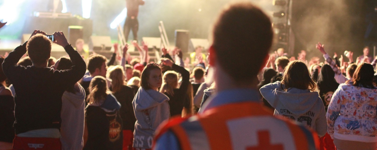 En hjelpekorpser med refleksvest med rødt kors på ryggen ser på en konsert med en stor menneskemengde foran scenen