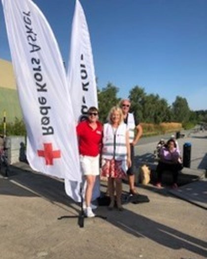 Tre personer fra Røde Kors, som står ved siden av to Røde Kors bannere. De står på en parkeringsplass på en dag med fint vær.