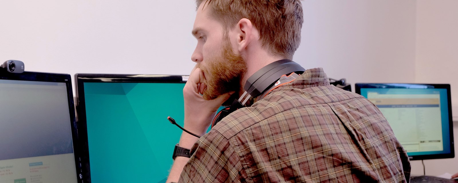 En mann sitter foran tre dataskjermer med et headset rundt nakken