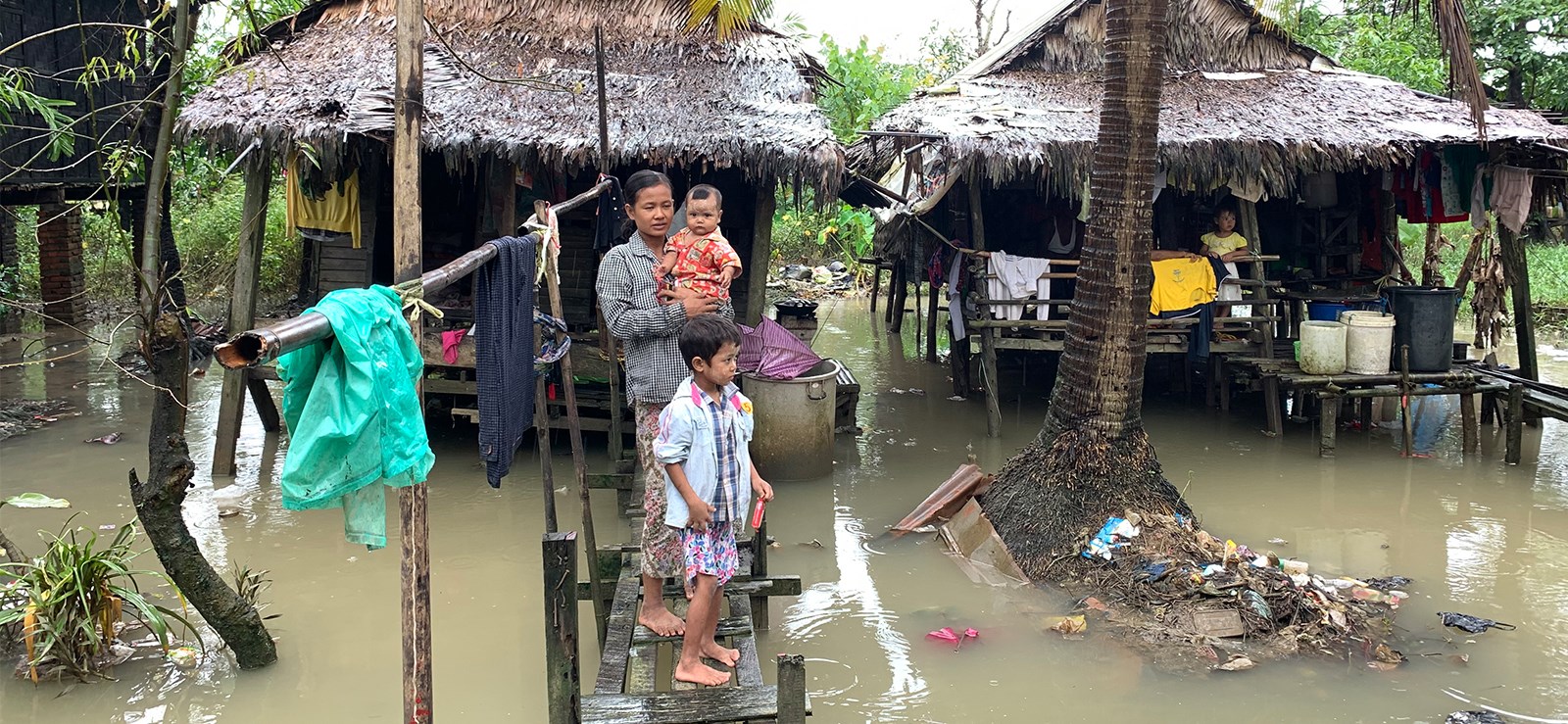 En kvinne og to barn står på en liten bro i en oversvømmet landsby