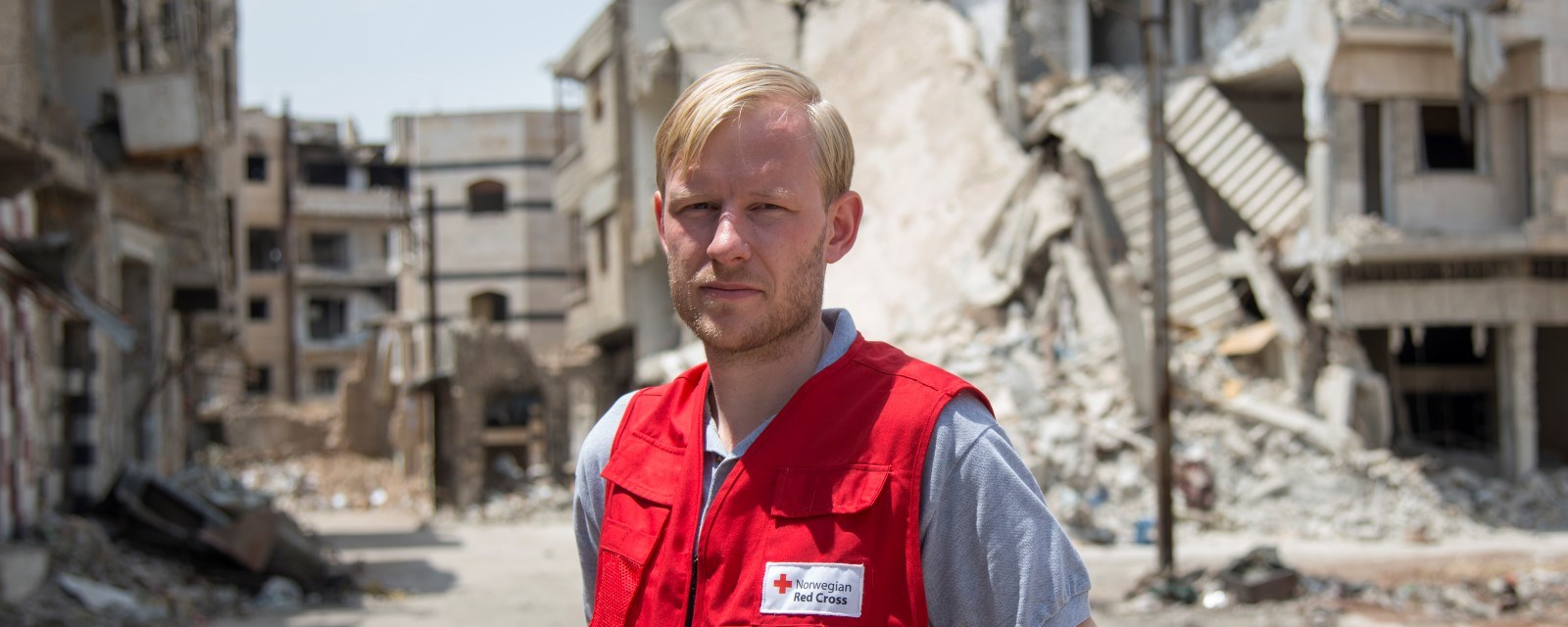 Ola Ulmo står blant ruiner og sønderbombede bygninger iført rød røde kors-vest