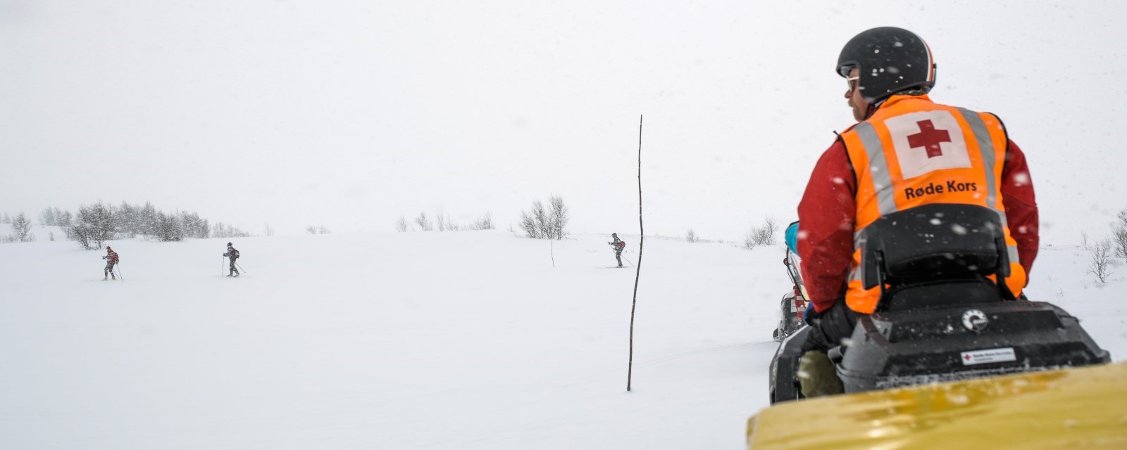 Skigåere i tett snødrev, med mann på Røde Kors-snøskuter som holder vakt