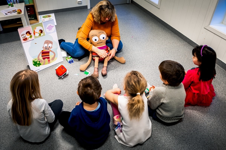 En gruppe med barn sitter og ser på en dame som holder en undervisningsdukke.