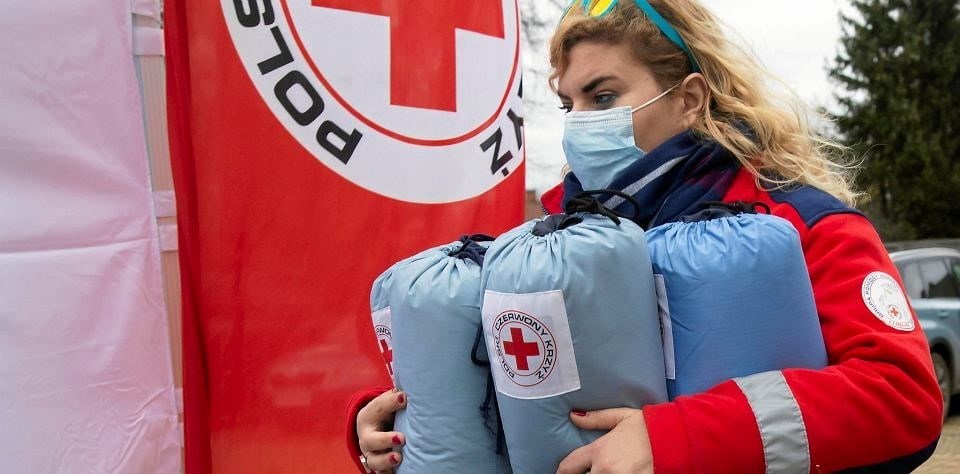 En kvinne med Røde Kors jakke og munnbind bærer noen pakker med Røde Kors logo på