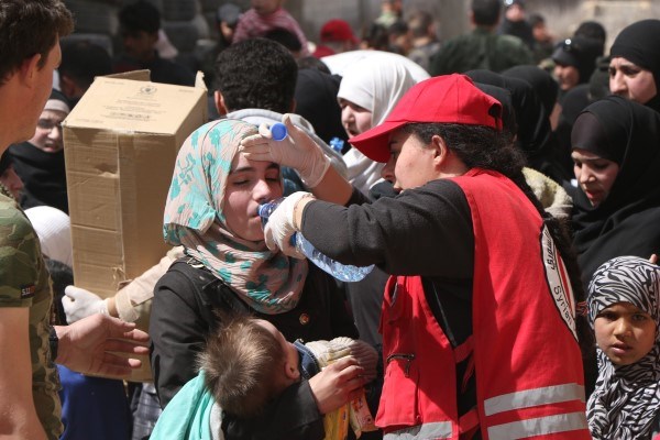 En ung kvinne med et lite barn i armene drikker vann fra en flaske holdt av en kvinnelig frivillig fra syrisk røde halvmåne.