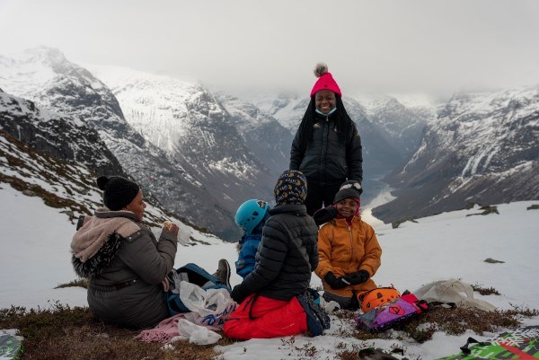 En voksen kvinne og tre barn sitter ute i naturen og spiser, ett barn står og smiler med utsikt over fjorden