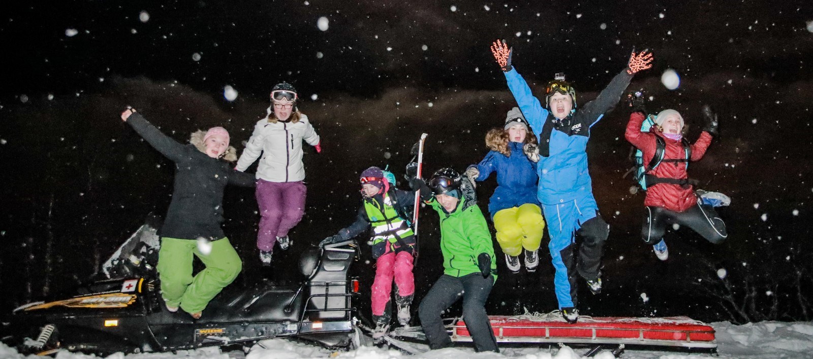 Fotografiet viser 7 ungdommer som hopper fra en snøskuter mens de ser i kameraet og strekker hendene i været.