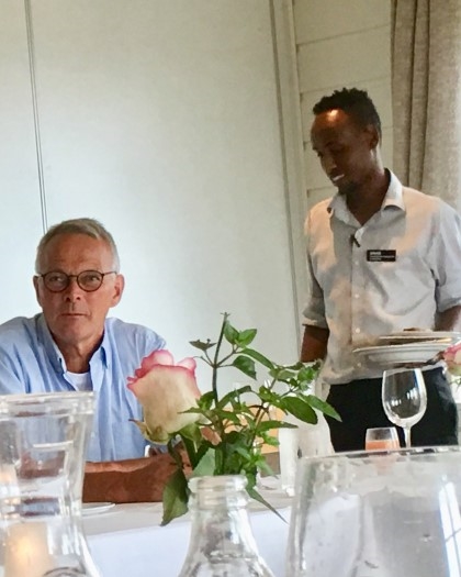 Bor tildekket med glass og blomster. En person sitter ved bordet og en servetør står bak og plukker opp talerkner.