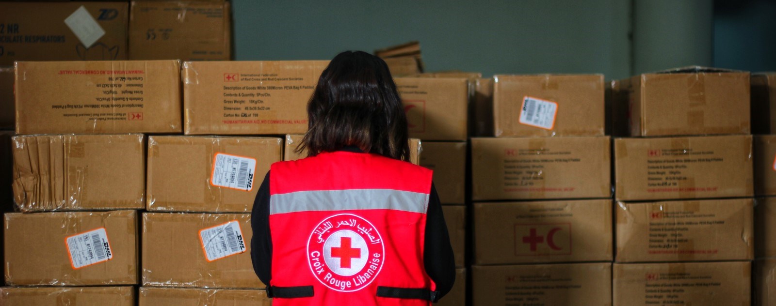 En kvinne med Røde Kors-vest står med rygget til og ser på en stabel med esker