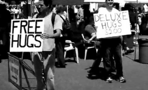 Gammelt bilde av to personer  hvor den ene holder en plakat hvor det står "Free hugs", mens den andres plakat sier "Hugs deluxe $2"