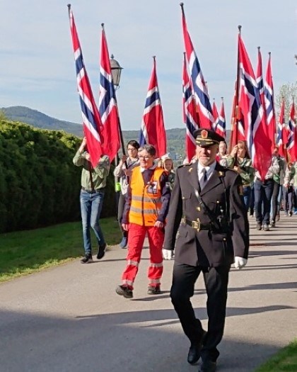 17 mai toget med æresmedlem av Asker Røde Kors foran. Speiderne går etter med store norske flag.