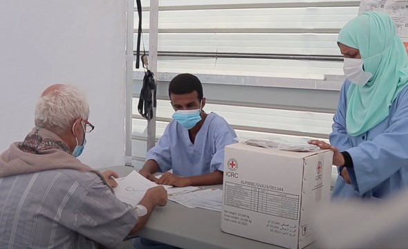 Mannlig pasient med munnbind sitter på mottaket til feltsykehuset sammen med to sykepleiere  i beskyttelsesutstyr som tar ham i mot