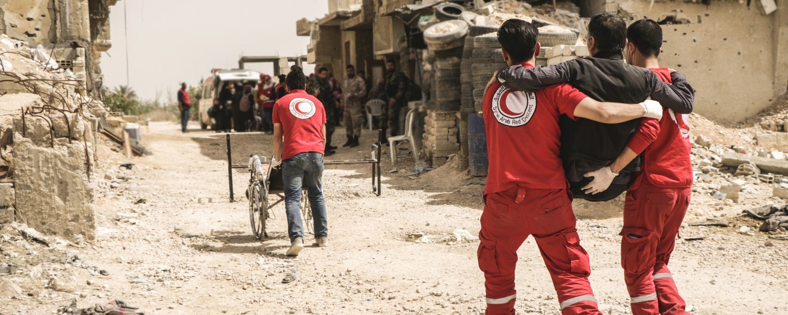 To frivillige fra Syrisk Røde Halvmåne bærer en skadet mann. En mannen frivillig går i front med en rullestol gjennom utbombede gater.