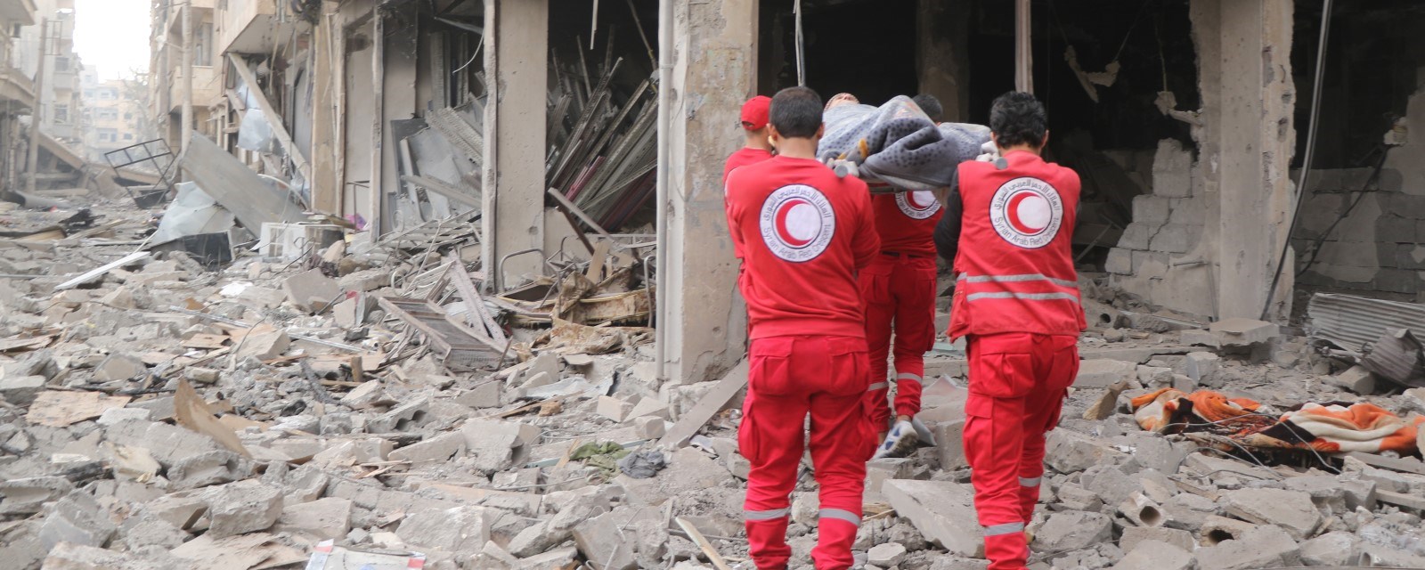 Fire frivillige fra Syrisk Røde Halvmånd bærer en person på båre i et utbombet område