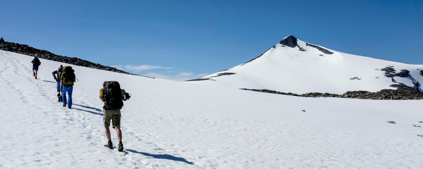 Bildet viser fire mennesker med ryggsekker på vei opp til en fjelltopp. De går på snø.