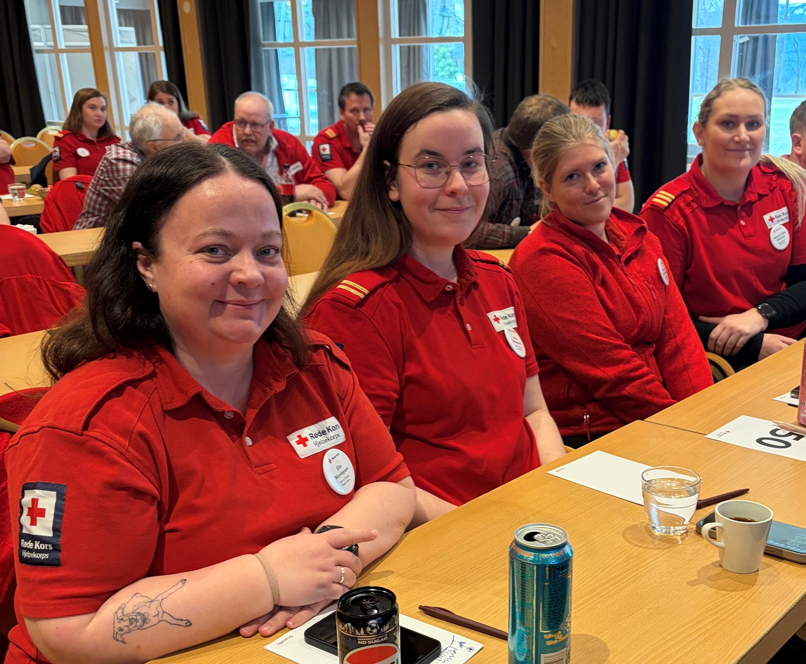 Fire kvinner og en mann i Røde Kors-klær på rekke i en møtesal.