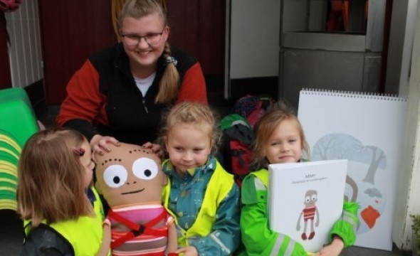 Dame med tre barnehage jenter kled i uteklær. De holder en dukke og en tenging av dukken.