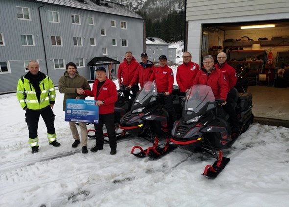 Røde Kors hjelpekorpsere med snøscootere står ute og smiler til kamera ved mottakelse av sjekk