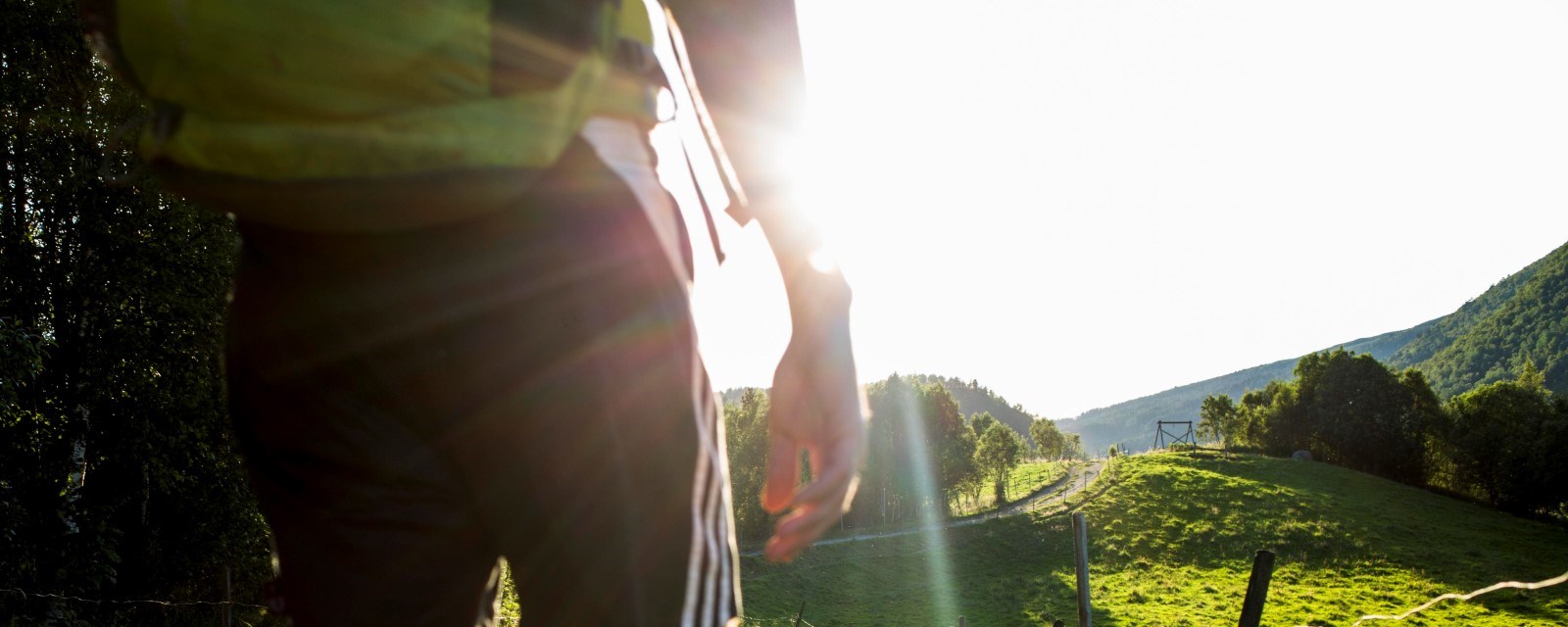 Bildet viser et menneske i kortbukse bakfra, som går i naturen i skinnende sol.