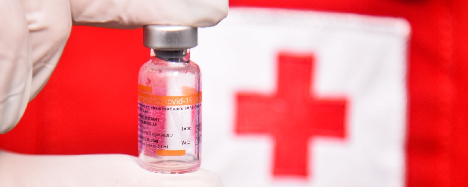 En liten flaske med koronavaksine holes av en person med Røde Kors uniform