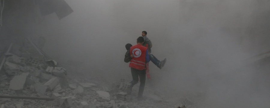 Mann i rød vest bærer en gutt i et område fylt av røyk og støv 