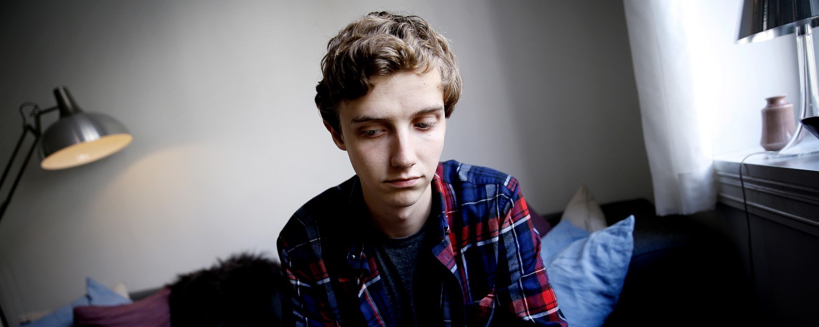 En ung mann sitter på en seng i et rom og ser ned i gulvet