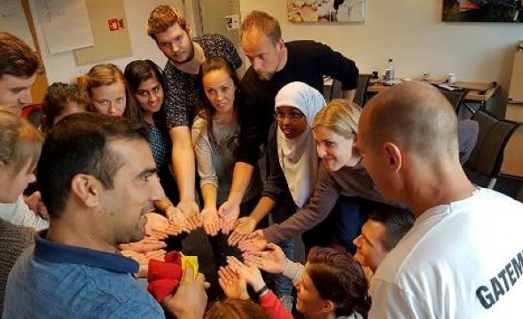 En gjeng med unge og voksne menn og kvinner, gutter og jenter står i ring og samler hendene sammen i senter.